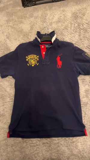 Mörkblå pikétröja Polo Ralph Lauren XL - Snygg mörkblå pikétröja från Polo Ralph Lauren i storlek XL. Tröjan har röda och gula broderade detaljer, vit rand på kragen och röd knappslå. Tillverkad i 100% bomull, med korta ärmar och klassisk polo-look. Perfekt för dig som gillar sportig och stilren design.