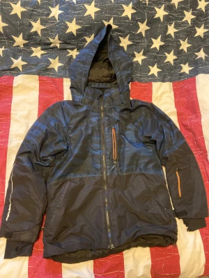 Blå camo vindjacka från Everest - Snygg blå vindjacka från Everest med camouflagemönster och orange detaljer på dragkedjorna. Jackan har huva, hel dragkedja framtill och flera praktiska fickor. Materialet är lätt och syntetiskt, perfekt för blåsiga dagar. Passformen är normal och jackan har långa ärmar. Jackan är till barn, det står inte vilken storlek det är på jackan men den passar till 140-150 eller 8-10 år gamla barn. På jackan finns det tecken på användning så den är alltså inte helt ny. 