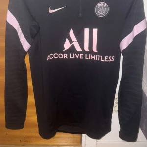 Svart PSG fotbollströja från Nike - Snygg svart PSG-tröja från Nike med rosa detaljer och texten 'Accor Live Limitless' på framsidan. Tröjan har långa ärmar och en dragkedja vid halsen. Nypris 850