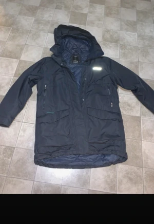 Mörkblå parkajacka från Didriksons - Säljer en mörkblå parkajacka från Didriksons med huva, flera fickor med dragkedja och tryckknappar. Jackan har quiltat foder och reflekterande logga på bröstet. Perfekt för kalla dagar och har en längre passform som täcker bra.