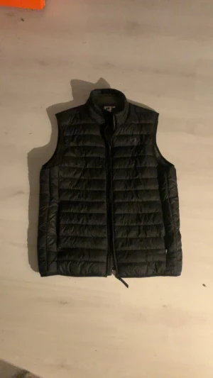 Svart dunväst med dragkedja från Hangten - Svart dunväst med horisontella quiltade sömmar och hög krage. Västen har en dragkedja framtill och är tillverkad i ett glansigt syntetmaterial. På bröstet finns en liten detalj med två silvriga fotavtryck. Perfekt för lager-på-lager under kyliga dagar.