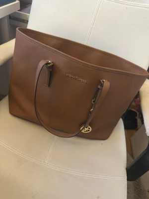 Brun axelväska från Michael Kors - Snygg brun axelväska från Michael Kors i skinn med guldfärgade detaljer och MK-logga. Väskan har flera fack, inklusive ett med dragkedja, och justerbara axelremmar. Perfekt storlek för att få plats med allt du behöver och stilren design.