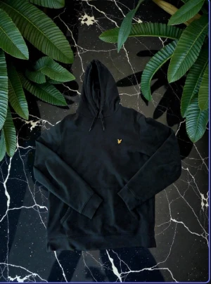 Grön hoodie från Lyle & Scott i M - Mörk grön hoodie från Lyle & Scott med klassisk gul fågel-logga broderad på bröstet. Tröjan har huva med snörning, lång ärm och en normal passform. Mjuk och skön bomullsmix som passar perfekt när du vill vara chill. Snygg och enkel design som funkar året runt.