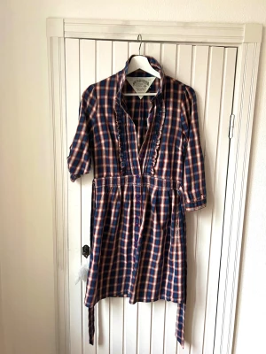 Kort rutig klänning / tunika från Hilfiger - Cool kort märkesklänning från T Hilfiger Denim med snygga krås-detaljer och fastsytt knytskärp. Knappar fram hela vägen, gör att man också kan bära den uppknäppt till byxor. Supermjukt bomullsmaterial i fräscht rutigt tyg med fantastisk färgkombo. Klassiskt plagg som kan stylas över jeans eller till strumpbyxor, och barbent på sommarhalvåret. Supersnygg till knähöga boots för den som gillar denim/ cowgirl stilen. 