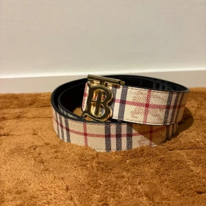 Rutig Burberry bälte med guldspänne - Säljer ett snyggt Burberry bälte med klassiskt rutigt mönster i beige, svart och rött. Bältet har ett exklusivt guldspänne med Burberrys ikoniska B-logga. Materialet är skinn med en lyxig känsla och passar perfekt till jeans eller kostymbyxor.