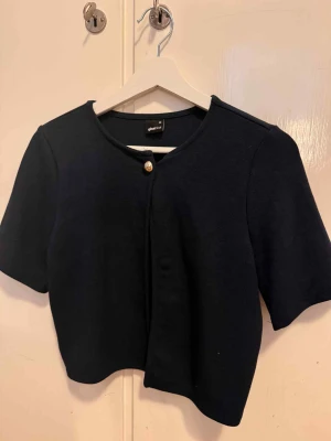 Mörkblå croppad kofta Gina Tricot - Snygg svart croppad cardigan/kofta jacka från Gina Tricot i storlek XS. Koftan har korta ärmar, rund halsringning och en guldfärgad knapp framtill. Perfekt för att lyfta din outfit med en stilren touch.