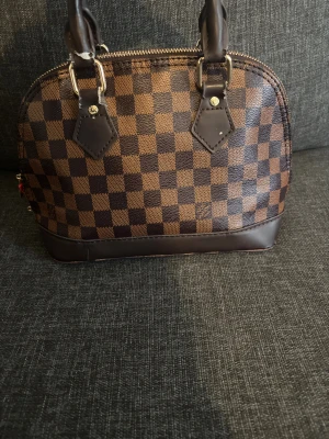 Brun  handväska  - Snygg handväska i klassiskt rutigt mönster med bruna och mörkbruna toner. Väskan har rundad form, två handtag, detaljer i mörkbrunt skinn och dragkedja upptill. Insidan är förstörd pga tvättning. 