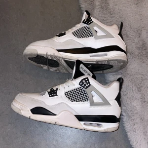 Nike Air Jordan 4 vit/svart/grå - Säljer ett par Nike Air Jordan 4 sneakers i vit, svart och grå färg. Klassisk siluett med meshpaneler, snörning och platt sula. Ikonisk Jumpman-logga på plösen och hälen. Skorna har detaljer i syntet och skinn, samt gråa och svarta inslag på sidorna och sulan.