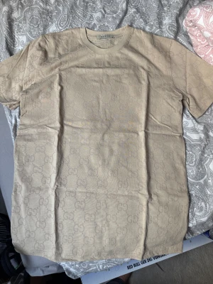 Beige Gucci t-shirt med GG-mönster - Snygg beige t-shirt från Gucci i bomull med diskret GG-monogram över hela tyget. Klassisk rund halsringning och korta ärmar. Perfekt för dig som gillar stilrena designer med lyxig känsla.