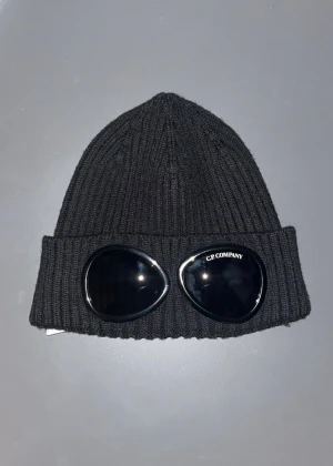 Svart Cp Company mössa - Säljer en svart ribbstickad mössa från C.P. Company med deras ikoniska goggle-detalj på uppviket. Mössan är tillverkad i mjukt stickat material och har en clean, urban look som sticker ut. Perfekt för dig som gillar streetwear och vill ha något unikt.
