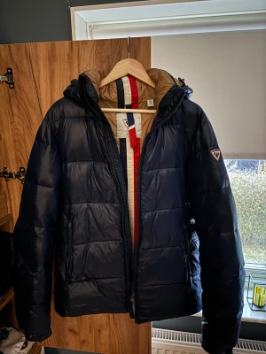 Mörkblå pufferjacka från Rossignol - Snygg mörkblå pufferjacka från Rossignol med beige foder och röd/blå/vita detaljer på insidan. Jackan har huva, dragkedja framtill och två sidofickor med dragkedja. Perfekt för kalla dagar eller skidåkning och har en Rossignol-logga på ärmen. Storlek M men passar även för L