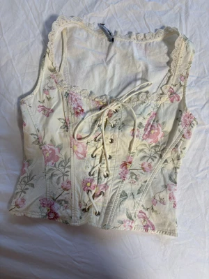 Blommig korsett från Gina Tricot - Superfin korsett från Gina Tricot i storlek 34. Toppen är cremevit med rosa och gröna blommor, har snörning framtill och spetsdetaljer vid halsringningen. Materialet är bomull och modellen är ärmlös med en feminin, figurnära passform. 100kr