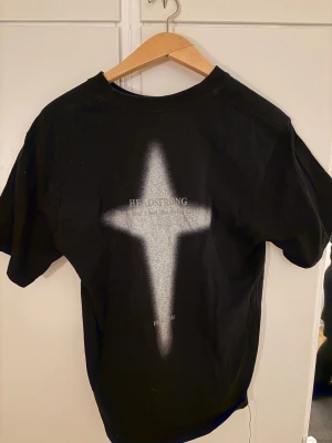 Svart t-shirt med vit korsdetalj - Svart t-shirt från Headstrong med vit korsliknande sprayad detalj på ryggen och texten 'FREEDOM' på bröstet. Klassisk rund hals och korta ärmar. Skön passform och tillverkad i mjuk bomull, perfekt för dig som gillar streetwear med edge.
