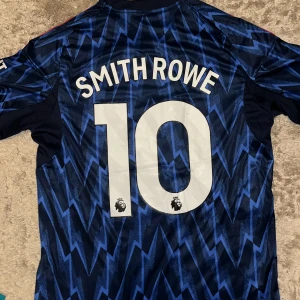 Arsenal Smith Rowe bortatröja Adidas L - Arsenal bortatröja med Smith Rowe 10 på ryggen. Mörkblå bas med ljusblå blixtmönster, röda Adidas-ränder på axlarna och klubbmärke på bröstet. Tillverkad i lätt och ventilerande material, perfekt för match eller träning.