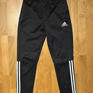 Svarta Adidas träningsbyxor 164 - Svarta träningsbyxor från Adidas med klassiska vita ränder längs benen och broderad logga på låret. Byxorna har elastisk midja och är gjorda i ett mjukt, syntetiskt material som passar perfekt för sport och chill.