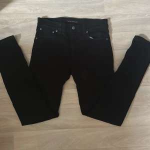 Svarta Nudie Jeans - Svarta jeans från Nudie Jeans med klassisk femficksdesign och raka ben. Jeansen har svarta sömmar, silverfärgade nitar och knapp, samt Nudie Jeans-logga på bakfickan. Tillverkade i jeansmaterial med en stilren och enkel look.