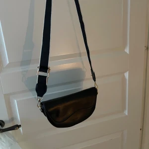 Svart axelväska i skinn med bred rem - Snygg svart axelväska i skinn med bred justerbar rem och guldfärgade detaljer. Väskan har ett lock med magnetknäppning och flera fack inuti, inklusive ett med dragkedja. Perfekt storlek för det viktigaste och stilren design som lyfter varje outfit.