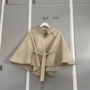 Beige cape-kappa från Missay - Stilren beige cape-kappa från Missay med snyggt bälte i midjan och dekorativ guldig knapp vid halsen. Jackan har en vid, svepande siluett och hög krage. Perfekt för dig som vill ha en elegant och trendig look under höst och vinter.