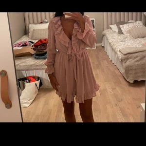 Rosa volangklänning med knytband - Supersöt kort rosa klänning med volanger längs ringningen och fållen. Klänningen har långa ärmar, v-ringning och ett knytband i midjan som ger en snygg siluett. Perfekt för dig som gillar romantisk och feminin stil. Nyskick!!!