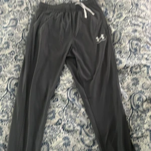 Grå träningsbyxor från Under Armour - Mörkgrå träningsbyxor från Under Armour med vit dragsko i midjan och logga på låret. Byxorna har elastisk midja och är gjorda i ett slätt, syntetiskt material som känns sportigt och bekvämt. Perfekta för gymmet eller chill hemma.