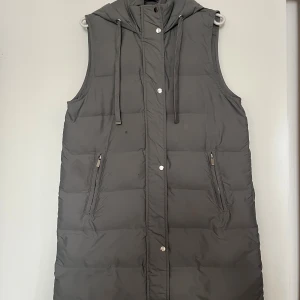 Grå lång dunväst med huva från ZARA - Lång, quiltad grå dunväst från ZARA med huva och dragsko. Västen har två snedställda fickor med dragkedja och stängs med både dragkedja och tryckknappar framtill. Perfekt för lager-på-lager och chill höst- eller vinterstil.