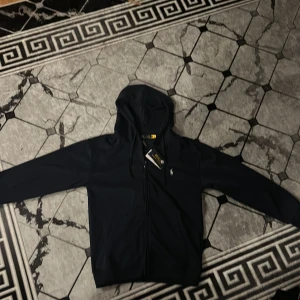 Svart hoodie från Polo Ralph Lauren - Svart hoodie från Polo Ralph Lauren med dragkedja och huva. Klassisk design med vit broderad logga på bröstet och snörning i huvan. Tillverkad i mjuk bomull, perfekt för en chill och stilren look.