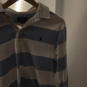 Randig pikétröja från Polo Ralph Lauren - Säljer en klassisk långärmad pikétröja från Polo Ralph Lauren i ljusblått och vitt. Tröjan har breda ränder, krage och knäppning framtill samt den ikoniska broderade loggan på bröstet. Perfekt för en preppy look!