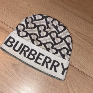 Beige Burberry mössa med logomönster - Snygg beige mössa från Burberry med ikoniskt monogrammönster i mörkgrått och stor logga framtill. Mössan är stickad och har uppvikt kant. Perfekt accessoar för att lyfta din vinterstil med lyxig känsla.