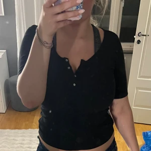 Svart topp med knappar från H&M - Svart topp från H&M med korta ärmar och djup v-ringning. Toppen har tre små knappar framtill och är figurnära i modellen. Perfekt att matcha med jeans eller kjol för en clean och enkel look.