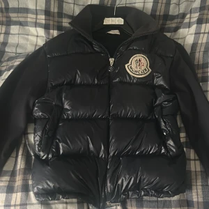 Svart pufferjacka från Moncler/ cardigan  - Säljer en svart pufferjacka från Moncler med glansig framsida och stora vadderade partier. Jackan har Moncler-logga på bröstet, dragkedja framtill och släta svarta ärmar. Perfekt för kalla dagar och riktigt snygg streetstil. 100% äkta nypris 10099kr. Finns en lite defekt sista bild inget som syns. Riktigt sällsynt jacka