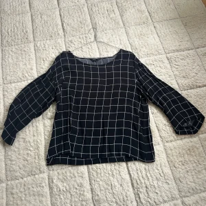 Svartvit rutig blus från MONKI - Svart blus från MONKI med vitt rutmönster och rund halsringning. Långärmad modell med lite lösare passform och raka ärmar. Tunt och mjukt material som känns skönt mot huden. Perfekt för dig som gillar stilrena och grafiska plagg.