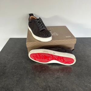 Christian Louboutin - Säljer ett par gråa sneakers från Christian Louboutin i mocka med ikoniska röda sulor och coola nitar på tån. Skorna har snörning och vit platt sula. Kommer med originalkartong och dustbag. Perfekta för dig som vill sticka ut med lyxig streetstyle.
