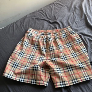Rutig Burberry shorts i bomull - Säljer ett par klassiska Burberry shorts/badshorts i storlek L med ikoniskt rutmönster i beige, svart, vit och röd. De har elastisk midja och är tillverkade i mjuk bomull med meshfoder. Perfekta för dig som gillar stilren design och vill sticka ut. 
