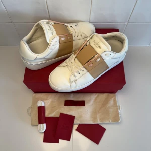 Valentino skor  - Snygga Valentino Garavani Open sneakers i vitt skinn med bred rosa rem över mitten och ikoniska nitar på hälen. Klassisk rund tå, platt sula och vita snören. Obs iget og tillkommer vid köp! Bara att höra av sig till oss vid minsta fundering 