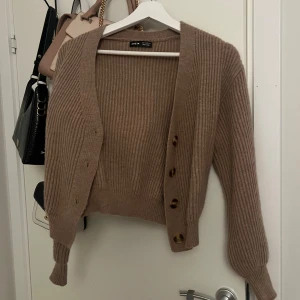 Beige ribbad kofta från Shein - Säljer en beige ribbad kofta från Shein med croppad passform. Koftan har långa ärmar, djup v-ringning och stora bruna knappar framtill. Perfekt för lager-på-lager och enkel att styla till olika outfits.