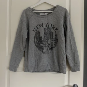 Grå sweatshirt med New York-tryck - Grå sweatshirt från ONLY ONE med stort New York-tryck framtill. Tröjan har rund halsringning, quiltade axelpartier och långa ärmar. Perfekt för en chill och avslappnad stil.