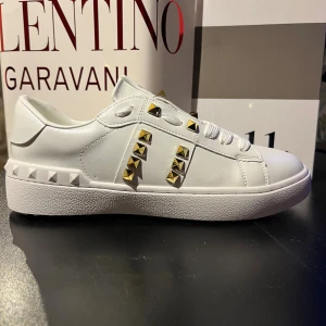 Valentino Rockstud Untitled sneakers - Snygga vita Valentino Rockstud Untitled sneakers i skinn med ikoniska guldfärgade nitar och chunky vit sula. Klassisk rund tå, snörning och coola detaljer längs sidorna och hälen. Perfekta för dig som vill sticka ut med exklusiv streetstyle.