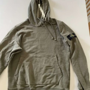 Grön hoodie från Stone Island - Säljer en grön hoodie från Stone Island med klassisk patch på ärmen. Tröjan har huva med snörning, magficka och långa ärmar. Tillverkad i mjuk bomull och har en relaxed passform. Perfekt för dig som gillar streetwear och vill ha en ikonisk look.