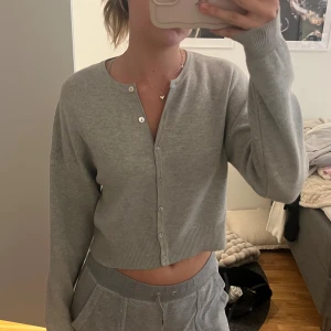 Grå croppad kofta med knappar - Grå croppad kofta med rund hals och långa ärmar. Tröjan har ribbade muddar och knäppning framtill med små knappar. Perfekt för lager-på-lager och chill dagar. Mjuk och skön känsla, passar till allt.