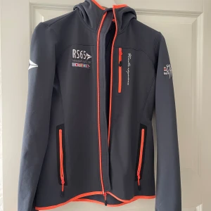 Svart vindjacka med orange detaljer - Svart vindjacka från Swedemount Nautic med orange dragkedjor och detaljer. Jackan har huva, tre fickor med dragkedja och broderade loggor på bröst och ärm. Perfekt för dig som gillar sportig och stilren look.