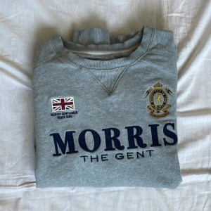 Grå sweatshirt från Morris  - Säljer en grå sweatshirt från Morris med broderad logga och text på bröstet. Tröjan har rund hals, långa ärmar och tryckt siffra på ena ärmen. Mjukt material och klassisk passform.