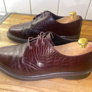 Bruna skinnloafers med krokoprägling - Snygga bruna loafers i skinn med krokodilpräglat mönster och rund tå. Skorna har snörning och en platt sula, vilket ger en klassisk och stilren look. Perfekta för dig som vill sticka ut med en unik detalj i outfiten.