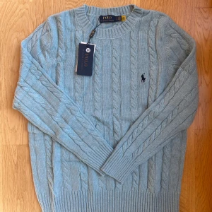 Ljusblå kabelstickad tröja Polo Ralph Lauren - Kabelstickad ljusblå tröja från Polo Ralph Lauren med klassisk rund halsringning och broderad logga på bröstet. Oanvänd, endast testad. Tröjan har långa ärmar och ribbade muddar vid ärmslut och nederkant. Perfekt för lager-på-lager under kyliga dagar. Passar till allt. Nypris: 2000kr