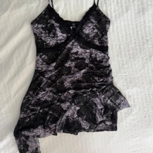 Klänning - Supersnygg kort klänning från weekday i lila och svart med blommigt spetsmönster. Klänningen har smala axelband, omlott-v-ringning och asymmetrisk nederkant med små shorts under.  Perfekt för dig som vill sticka ut med en edgy och feminin look.