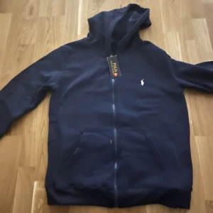 Mörkblå hoodie från Polo Ralph Lauren - Snygg mörkblå hoodie från Polo Ralph Lauren med klassisk vit logga på bröstet. Tröjan har huva med snörning, dragkedja framtill och två fickor. Perfekt för dig som gillar stilren och bekväm streetwear.