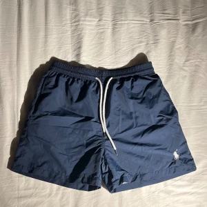 Blå badshorts från Polo Ralph Lauren - Snygga marinblå badshorts från Polo Ralph Lauren med vit broderad logga på benet. Shortsen har elastisk midja med vit snörning, innerbyxa i mesh och en bakficka med lock. Perfekta för stranden eller poolen. Storlek L men sitter som M.