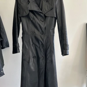 Svart lång skinn trenchcoat - Säljer en svart lång trenchcoat i skinn med bred krage, dubbelknäppning och snygga detaljer som bälte i midjan och stora fickor. Jackan har långa ärmar och en cool, oversized siluett som ger riktigt edgy vibes. Perfekt för dig som vill sticka ut med en statement piece. Old leather jacket🦇