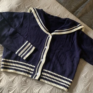 Marinblå kofta med sjömanskrage  - Snygg marinblå kofta med vitrandiga detaljer på krage, muddar och nederkant. Koftan har knappar framtill och en cool sailor-inspirerad krage. Perfekt för dig som gillar retrovibbar och vill sticka ut med en unik look.