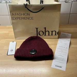 Vinröd merinoullsmössa från C.P. Company - Snygg vinröd ribbstickad mössa från C.P. Company i mjuk merinoull. Klassisk beanie-modell med uppvikt kant och rund svart logotypdetalj framtill. Perfekt för dig som vill ha en stilren och varm accessoar med streetkänsla. Storleken är one size så den passar alla huvudformer och storlekar. Det är bara att höra av sig vid frågor och funderingar 😀