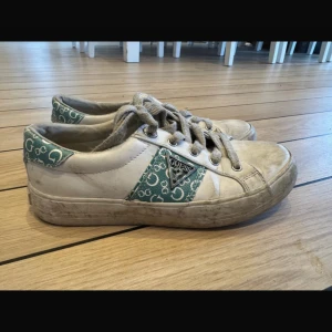 Guess vita sneakers med grönt mönster - Snygga vita sneakers från Guess med gröna detaljer och logomönster på sidor och häl. Skorna har snörning, platt sula och rund tå. Materialet är skinn och textil, med metallfärgad Guess-logga på sidan. Perfekta för en trendig vardagslook. Storlek 38